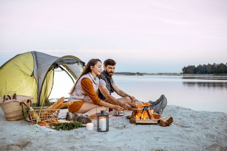 Young and cheerful couple sitting at the fireplace, cooking sausages, having a picnic at the campsite on the beach in the eveningの写真素材