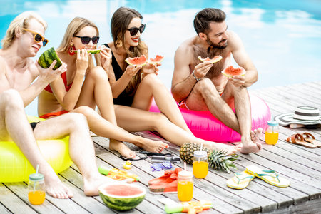 Group of a happy friends in swimwear eating watermelon while sitting together and having fun during...