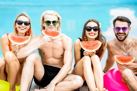 Portrait of a group of a happy friends in swimwear eating watermelon while sitting together on the swimming pool in the summerの写真素材
