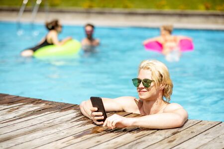 Man using smart phone while resting with friends in the swimming pool outdoors during the summertimeの写真素材