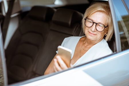 Senior business woman using smart phone while sitting on the back seat of the modern car. Concept of a business life on retirementの写真素材
