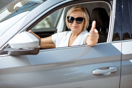 Portrait of a happy senior woman driver looking out the car window and showing ok sign. Concept of an active people during retirement ageの写真素材