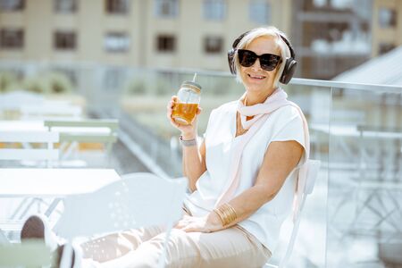 Portrait of a stylish senior woman dressed in white enjoying music while sitting with summer drink on the terrace outdoorsの写真素材