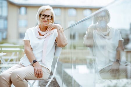 Lifestyle portrait of an elegant senior woman dressed in white sitting on the cafe terrace outdoors. Concept of an active lifestyle on retirementの写真素材