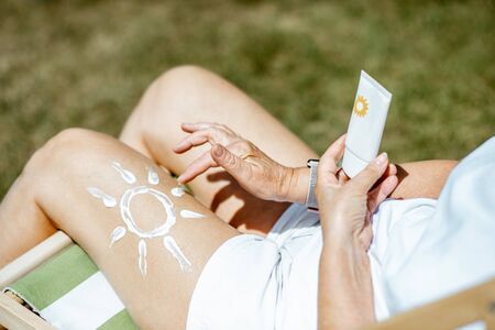 Senior woman with sun shape on her leg made of sunscreen lotion, holding a tube with cream while resting outdoorsの写真素材