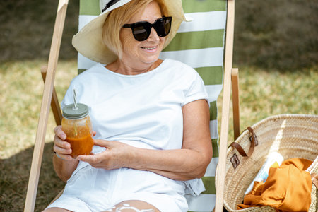 Beautiful senior woman in white clothes lying with juice on the sunbed, relaxing in the park. Concept of rest and carefree life on retirementの写真素材
