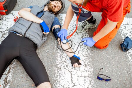Medic applying first aid to the injured cyclist lying on the pedestrian crossing after the road accident, doctor measuring pressureの写真素材