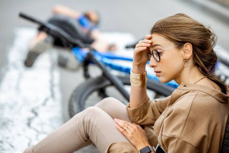 Sad woman sitting near the car, worried of injured man lying on the pedestrian crossing after the road accidentの写真素材