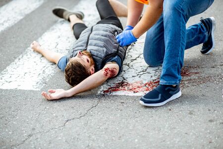 Man applying first aid to the injured bleeding person, wearing tourniquet on the arm after the road accident on the pedestrian crossing