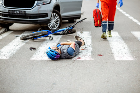 Road accident with injured cyclist lying on the pedestrian crossing, medic going to apply first aidの写真素材