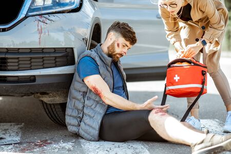 Driver hurrying with first aid kit to help injured man with bleeding wounds sitting near the car after the road accidentの写真素材