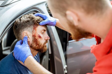 Ambulance worker examining facial injuries of a man sitting near the car after the road accident, providing emergency medical assistanceの写真素材