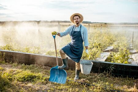 Well-dressed farmer with working tools standing on a farmland with automatic watering on a farm during the sunsetの写真素材