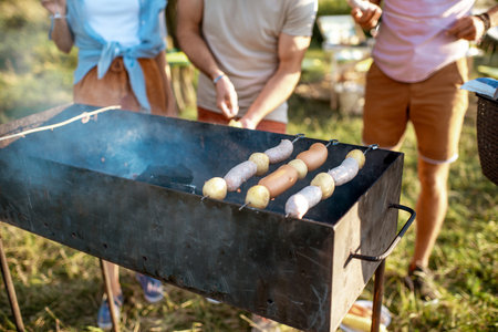 Friends cooking sausages and potatoes on a barbecue outdoors, close-up view