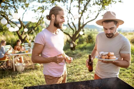 Two male friends cooking food on a barbecue in the beautiful garden during festive lunch with girlfriends on a sunny afternoonの写真素材