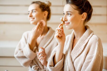 Young women taking care of their skin, making facial massage in the SPA, sitting together in bathrobesの写真素材