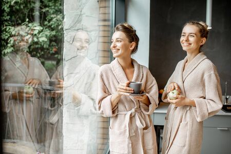 Portrait of a two young women in bathrobes standing together on the kitchen near the window, relaxing after the SPA proceduresの写真素材