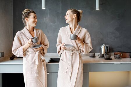Two young women in bathrobes standing together on the kitchen of the modern SPA complex or home, drinking coffee after the SPA proceduresの写真素材
