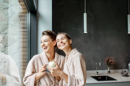 Portrait of a two young women in bathrobes standing together on the kitchen near the window, relaxing after the SPA proceduresの写真素材
