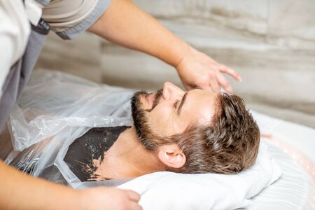 Man during a mud wrapping with special black mud, lying in the spa salon. Concept of a Detox mud body wrapsの写真素材