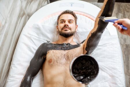 Man during a mud wrapping with special black mud, lying in the spa salon. Concept of a Detox mud body wrapsの写真素材