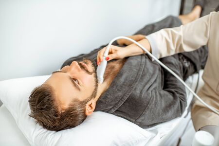 Men examining his thyroid with ultrasound sensor, lying in bathrobe on the couch at the medical officeの写真素材