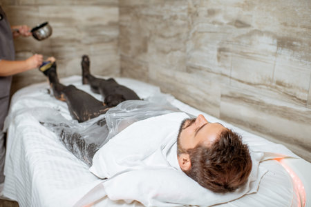 Man during a mud wrapping with special black mud, lying in the spa salon. Concept of a Detox mud...