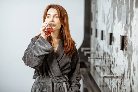Beautiful woman in bathrobe drinking mineral water with wine extract at the pump-room while resting at the luxury resortの写真素材