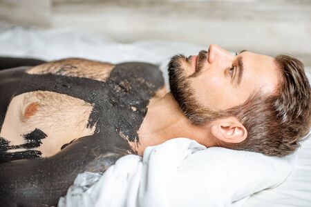 Man during a mud wrapping with special black mud, lying in the spa salon. Concept of a Detox mud body wrapsの写真素材