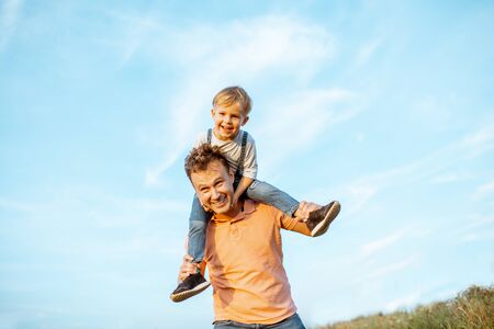 Portrait of a happy father with young son riding on the shoulders on the blue sky background outdoors. Concept of a happy family on a summer activityの写真素材