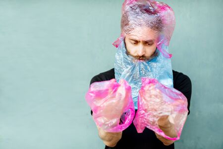 Portrait of confused man with plastic bags on hands and head standing on the green background. Concept of non-recyclable plastic pollutionの写真素材