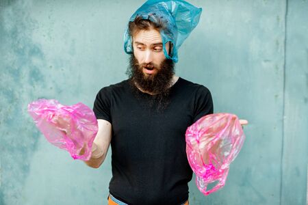 Portrait of confused man with plastic bags on hands and head standing on the green background. Concept of non-recyclable plastic pollutionの写真素材