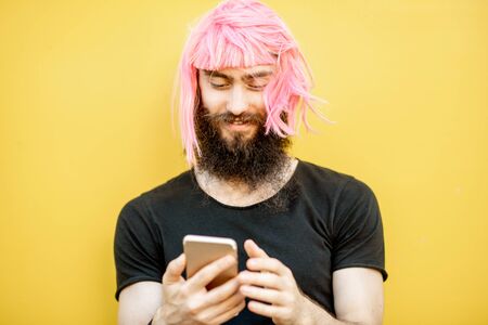 Funny portrait of a stylish man with beard and long color hair usning smart phone on the yellow backgroundの写真素材