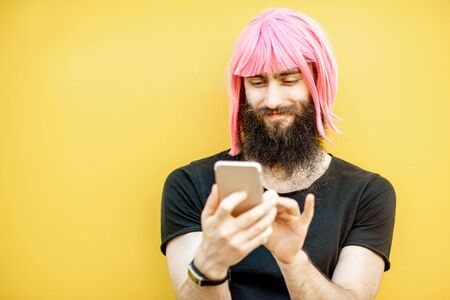 Funny portrait of a stylish man with beard and long color hair usning smart phone on the yellow backgroundの写真素材
