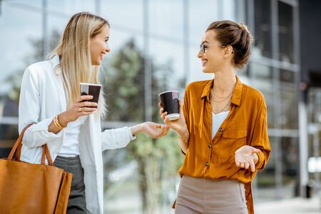 Two young businesswomen talking near the office building, having a small talk during the coffee break outdoors.の写真素材