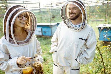 Two young beekepers in protective uniform working on a small apiary farm with wooden beehives on the backgroundの写真素材