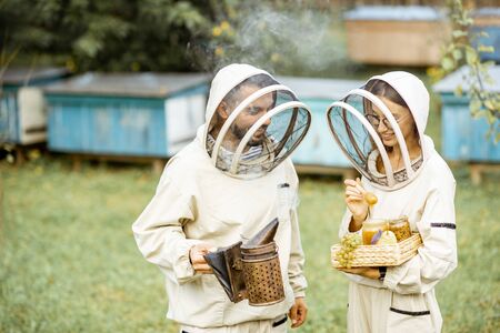 Two beekepers in protective uniform standing together with beesmoker and honey, tasting fresh product on the apiaryの写真素材