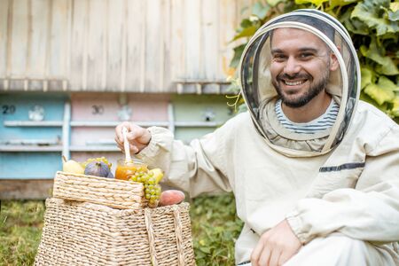 Portrait of a cheerful beekeeper in protective uniform with honey products and sweet fruits on the apiary with beehives on the backgroundの写真素材