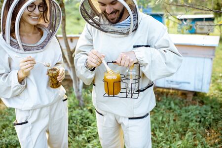 Two beekepers in protective uniform standing together with honey in the jar, tasting fresh product on the apiary outdoorsの写真素材