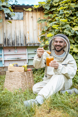 Portrait of a cheerful beekeeper in protective uniform with honey products and sweet fruits on the apiary with beehives on the backgroundの写真素材