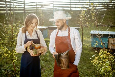 Potrait of a man and woman farmers in aprons standing with beesmoker, fruits and honey on the apiary with beehives on the background on a sunny eveningの写真素材