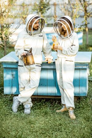 Two beekeepers in protective uniform standing together near the wooden beehives on a small traditional apiary. Concept of beekeeping and small farmingの写真素材