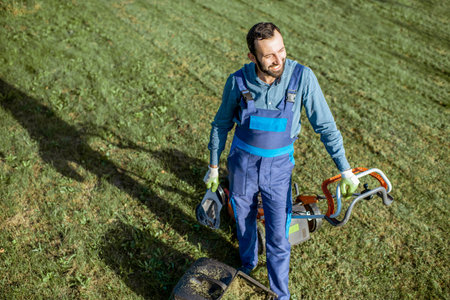 Portrait of a professional gardener in protective workwear resting while cutting grass with gasoline...