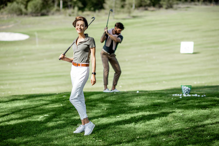 Portrait of an elegant young woman standing with golf putter and friends playing golf on the backgroundの写真素材