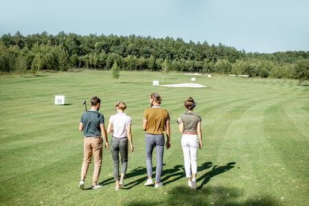 Young and elegant friends walking with golf equipment while playing golf on the beautiful course on a sunny day, view from the backsideの写真素材