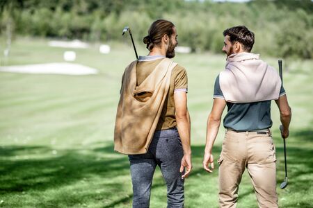 Two male best friends walking with golf equipment on beautiful playing course, talking and having fun during a game, rear viewの写真素材