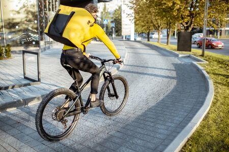 Young bearded courier delivering food with a yellow thermal backpack, riding a bicycle in the city. Food delivery service conceptの写真素材