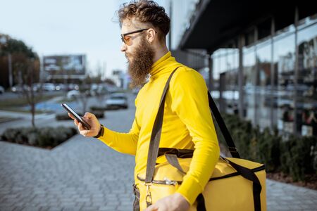 Young courier delivering food with thermal bag, checking order on a smart phone while standing on the city street. Delivery on foot conceptの写真素材