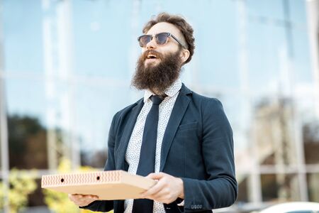 Portrait of a happy bearded businessman dressed in the suit going for lunch with pizza on the office building background outdoorsの写真素材