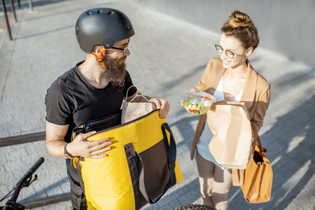 Male courier delivering food on a bicycle, giving bag with fresh takeaway lunch to a young business...